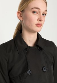 Crni trench kaput s reverima, dvostrukim kopčanjem s gumbe i glatkim teksturiranim materijalom. Osvijetljen velikim gumbima.