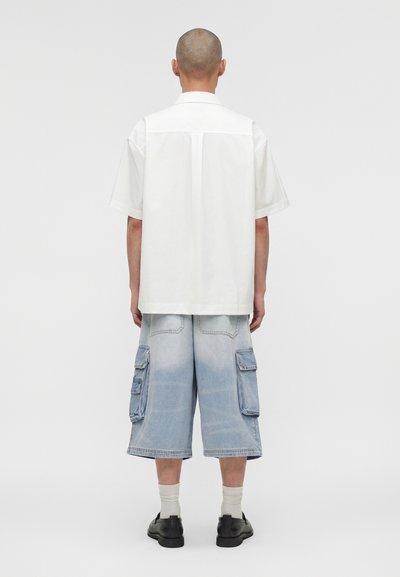 Άτομο με ξυρισμένο κεφάλι που φοράει λευκό κοντομάνικο πουκάμισο, ανοιχτό μπλε cargo σορτς μέχρι το γόνατο, άσπρες κάλτσες και μαύρα παπούτσια, στέκεται με την πλάτη γυρισμένη.
