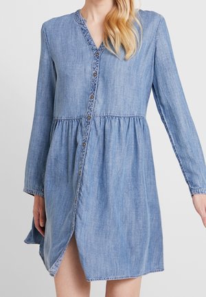 Vrouw draagt een lichtblauwe denimjurk met lange mouwen, knopen aan de voorkant en een geplooide taille, staand tegen een effen achtergrond.