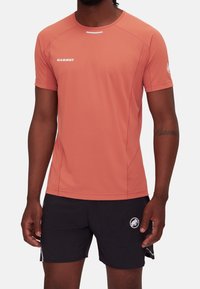 Man wearing a coral short-sleeve Mammut shirt and black athletic shorts with white Mammut logos on chest, sleeve, and leg.