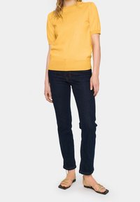 Žlutý pletený top s krátkým rukávem a kulatým výstřihem, sladěný s tmavě modrými džíny. Modelka má na sobě světle zelené sandály na tenkých páscích se čtveratou špičkou.