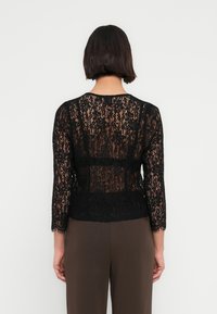 Μαύρο τοπ από δαντέλα με μανίκια τριών τετάρτων, με λουλουδένιο σχέδιο και διαφανή υφή, φορεμένο πάνω από καφέ παντελόνια.