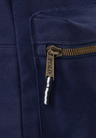 Marine canvas tas met een messing rits, voorzien van een label met "POLO", en een zwart-witte stoffen trekband. Textuur oppervlak.