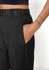 Mustat shortsit, jotka on valmistettu kevyestä materiaalista ja joissa on vetoketjullinen tasku sekä kuminauhavyötärö, tiiviillä verkkomaista rakennetta oleva.