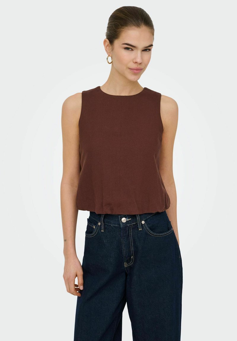 Femme aux cheveux noirs attachés en chignon, portant un haut marron sans manches et un jean bleu foncé taille haute, debout devant un fond clair.