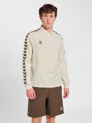 Hombre con camisa polo de manga larga color crema con rayas en forma de cheurón marrones en las mangas y pantalones cortos marrones con logo en la pierna izquierda sobre fondo blanco.