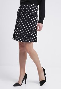 Falda negra con lunares blancos, de longitud a la rodilla, hecha de tejido suave. Presenta una cintura ajustada y cae suavemente en el dobladillo. Tacones negros de punta.