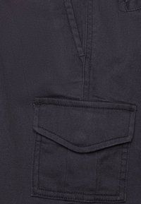 Tessuto blu scuro con una superficie texture, caratterizzato da una grande tasca con patta e dettagli di cucitura lungo i bordi.