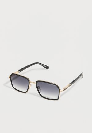 Square-shaped sunglasses with gradient dark lenses, gold metal frame, and black arms, placed on a white background.