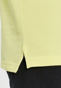 Camisa amarilla de manga corta con un tejido texturizado y aberturas en los lados del dobladillo. La camisa presenta costuras limpias y un corte holgado.