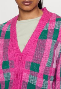 Femme portant un cardigan en maille à carreaux roses et verts sur un haut beige clair à col rond, visible du cou jusqu’au milieu du torse.