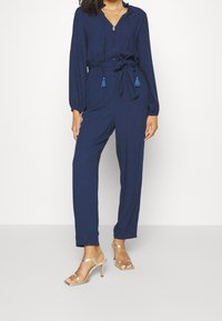 Navy blue jumpsuit with long sleeves, a zipper front, drawstring waist, and tassel accents. Smooth fabric with subtle texture.
