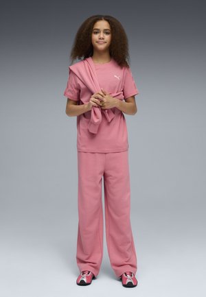 Joven con cabello rizado vistiendo un conjunto rosa de Puma, sosteniendo una sudadera rosa a juego, posando sobre un fondo gris.