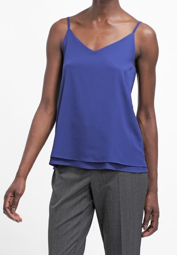 Personne portant un haut bleu sans manches à col en V et un pantalon gris, debout devant un fond blanc uni.