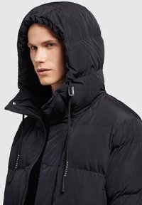 Fekete pufi dzseki magas gallérral és állítható kapucnival. Simma, fényes felülettel rendelkezik, fekete meghúzókkal, márkás díszítésekkel.