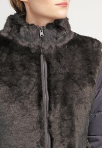 Kamizelka z sztucznego futra w ciemnoszarym kolorze z wysokim kołnierzem, zamek błyskawiczny i gładkie krawędzie. Posiada pluszową fakturę oraz kontrastowy materiał na rękawach.