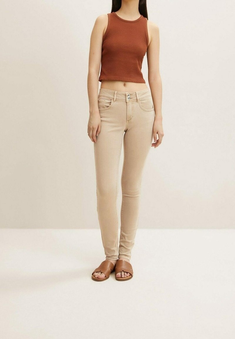 Femme portant un top court marron sans manches, un jean skinny beige et des sandales marron à bout ouvert, debout devant un fond clair.
