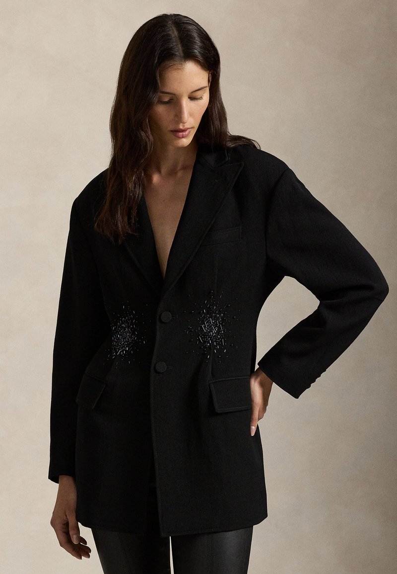 Black blazer with a fitted design, two front pockets, and subtle beaded detailing on the chest. Made of textured fabric and single-breasted.