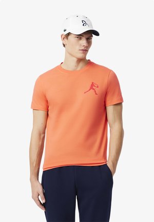 Orange cotton t-shirt med korte ærmer og rund halsudskæring, med en pink silhuetlogo på venstre bryst. Kombineret med marineblå bukser.