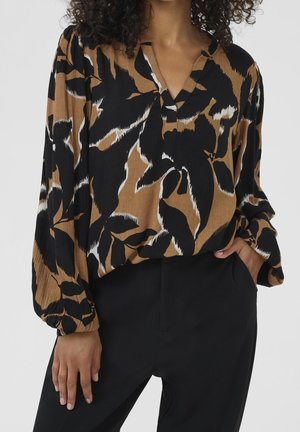 Mujer con blusa de manga larga con patrones abstractos de hojas en negro, marrón y blanco, y pantalones negros, con la mano en el bolsillo.