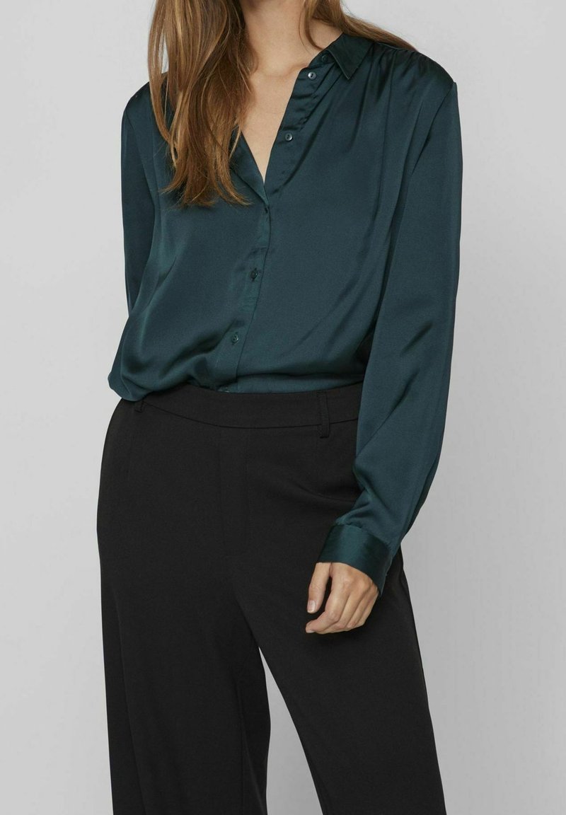 Blouse en satin vert foncé à manches longues, col à revers et fermeture par boutons, associée à un pantalon noir ajusté. Texture lisse, design épuré.