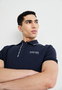 Jūras spēku stila polo krekls ar īsām piedurknēm, divu pogu aizdare un balti akcenti apkaklēs. Piedāvā Red Bull Racing logo detaļu.