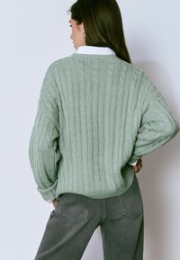 Mujer con pelo largo castaño que lleva un suéter de punto de cable verde claro sobre una camisa blanca y pantalones gris oscuro, de espaldas con las manos en las caderas.