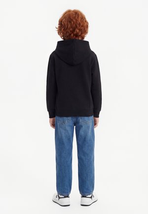 Enfant aux cheveux roux bouclés portant un sweat à capuche noir, un jean bleu et des baskets blanches, debout de dos sur un fond blanc.