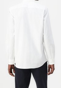 Vit långärmad skjorta med krage, med knäppning vid ärmsluten och rak fåll. Tyget verkar slätt och lätt.