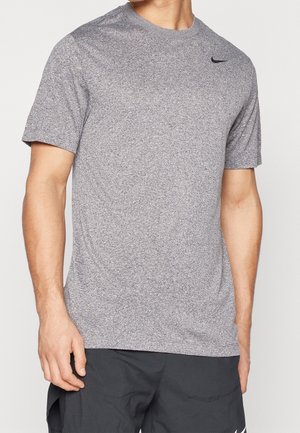 Hombre con camiseta deportiva gris de manga corta Nike y pantalones cortos negros, de pie frente a un fondo claro y liso.