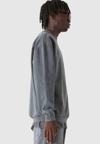 Szürke kapucnis pulóver puha anyagból, laza szabással, bordázott mandzsettákkal és bokákkal, valamint látható varrási részletekkel. Réteges szürke öltözetben viselve.