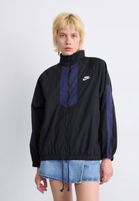 Črna vetrovka z vijolično ploščno, z zadrgo spredaj, visokim ovratnikom in nastavljivim robom. Nike logotip v belem na prsih.