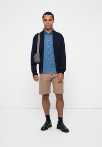 Marine cardigan, blå denimskjorte, beige ribbestrikkede shorts, svarte slip-on-sko, grå crossover-veske, står foran en ensfarget hvit bakgrunn.