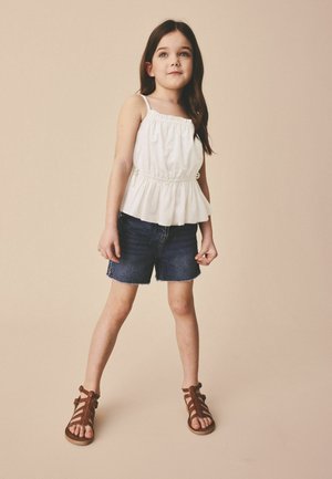 Noor tüdruk seisab käed külgedel, kandmas valget varrukateta toppi, teksapükse ja pruune rihmikuga sandaale ühevärvilise beeži tausta ees.