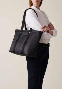 Borsa a tracolla nera con texture e manici con logo verticale, caratterizzata da una forma strutturata e dettagli metallici. Abbinata a una camicia bianca con bottoni.