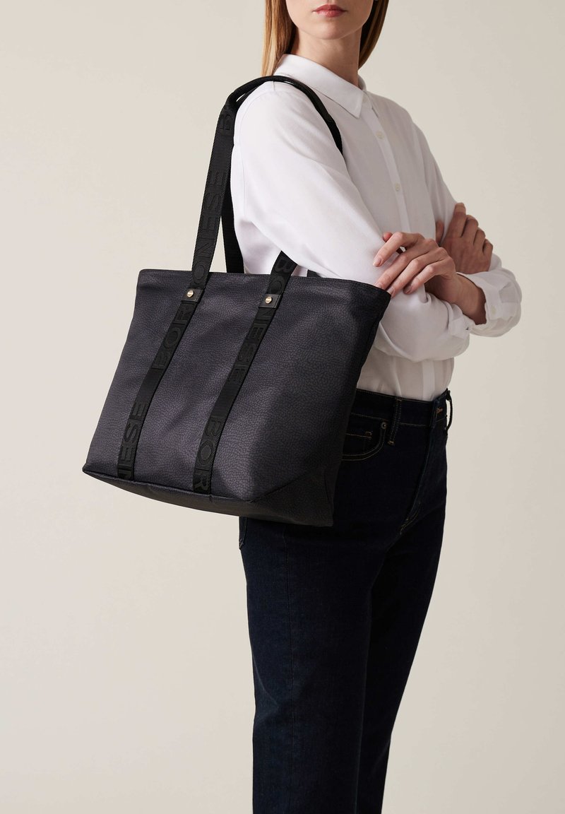 Borsa a tracolla nera con texture e manici con logo verticale, caratterizzata da una forma strutturata e dettagli metallici. Abbinata a una camicia bianca con bottoni.