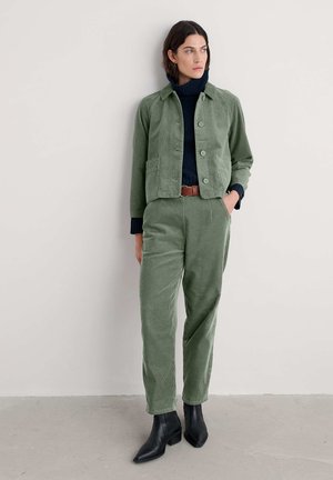 Vrouw in een olijfgroene corduroy jas en broek, marineblauwe coltrui, bruine riem en zwarte enkellaarsjes, staand tegen een lichte muur.