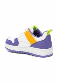 Zapatilla blanca con acentos morados, cinta lateral naranja y cuello verde. Presenta una superficie texturizada y una suela de goma resistente.