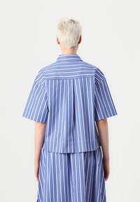 Camicetta a maniche corte di colore blu chiaro con righe verticali bianche. Presenta un colletto, una piega sul retro e un taglio rilassato. Realizzata in tessuto leggero.