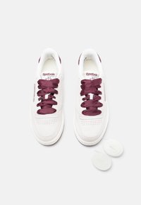 Białe zamszowe sneakersy z bordowymi satynowymi sznurówkami, z perforowanymi detalami na palcach oraz logo Reebok na języku. W zestawie dodatkowe sznurówki.