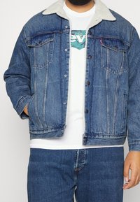 Man med blå denimjacka och vit fårost krage, vit grafisk t-shirt och blå jeans, stående med ena handen i fickan.