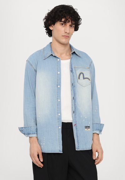 Camicia in denim azzurro chiaro con colletto, tasca anteriore con design curvo, chiusura a bottoni e dettagli sfrangiati sull'orlo inferiore.