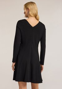 Schwarzes Langarmkleid mit V-Ausschnitt auf der Rückseite, tailliertem Oberteil, weit schwingendem Rock und dezentem Nahtdesign. Glatte Textur und mittlere Oberschenkel-Länge.