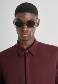 Gafas de sol con un marco gris transparente y lentes oscuros, combinadas con una camisa de color burdeos oscuro con cuello. Líneas limpias y diseño angular.