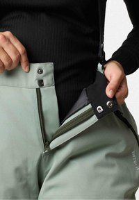 Mains tenant un pantalon vert ouvert à glissière avec un bouton-pression noir, montrant la doublure intérieure en tissu noir et les bretelles attachées.