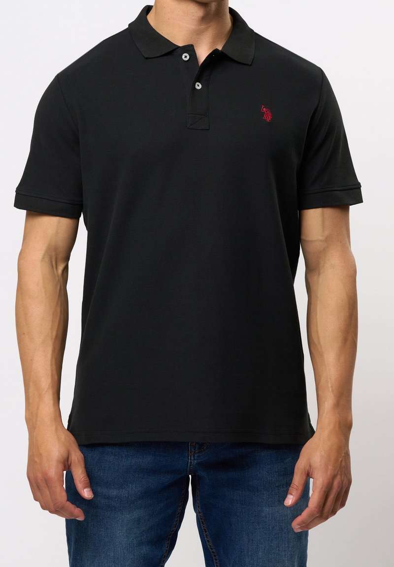 Homme portant un polo noir à manches courtes avec un logo rouge sur la poitrine gauche et un jean bleu, debout devant un fond uni.