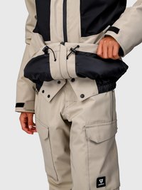 Jachetă bej și neagră cu talie ajustabilă și pantaloni cargo cu buzunare cu clapetă, prezentată pe o persoană care își ajustează jacheta.