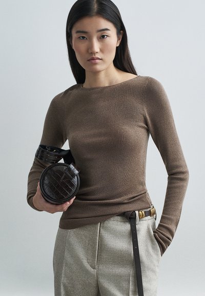 Bruine geribbelde longsleeve top gedragen met lichtgrijze broek. Het model houdt een ronde donkerbruine tas met een gestructureerde afwerking en een zwarte riem met gouden accenten vast.