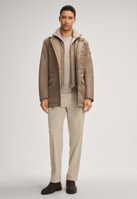 Braune wasserdichte Jacke mit Schaffellkragen, kombiniert mit einem hellbeigen Pullover und beigen Hosen. Das Modell steht in dunklen Stiefeln.