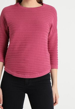 Femme portant un pull rose texturé à manches longues avec un ourlet arrondi, associé à un pantalon noir, sur un fond uni.